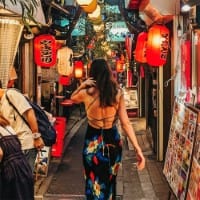 A woman dressed in a colourful dress with open back strolls through a narrow bustling market street of Tokyo. Products are displayed outside shops to tempt passing patrons as they walk underneath chinese laterns
